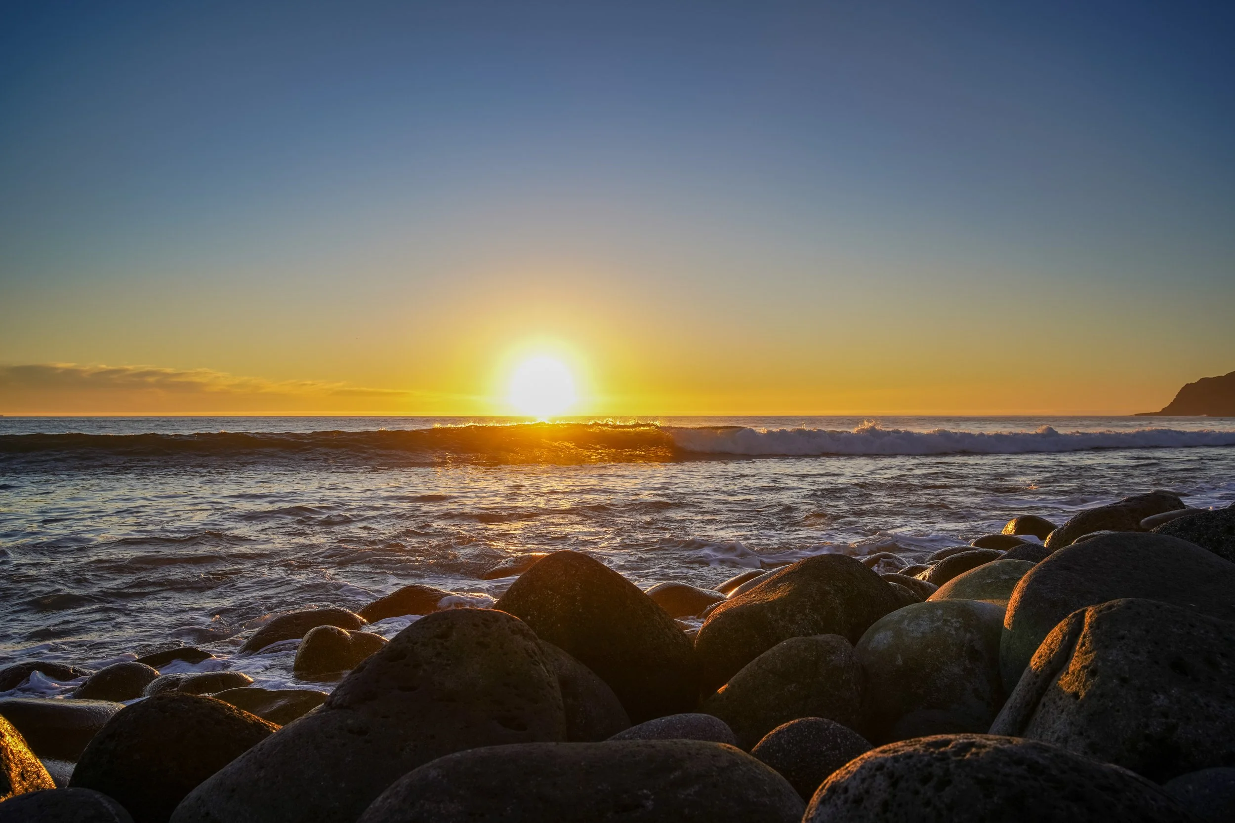 Sunset over the ocean with waves breaking on a rocky shoreline.
