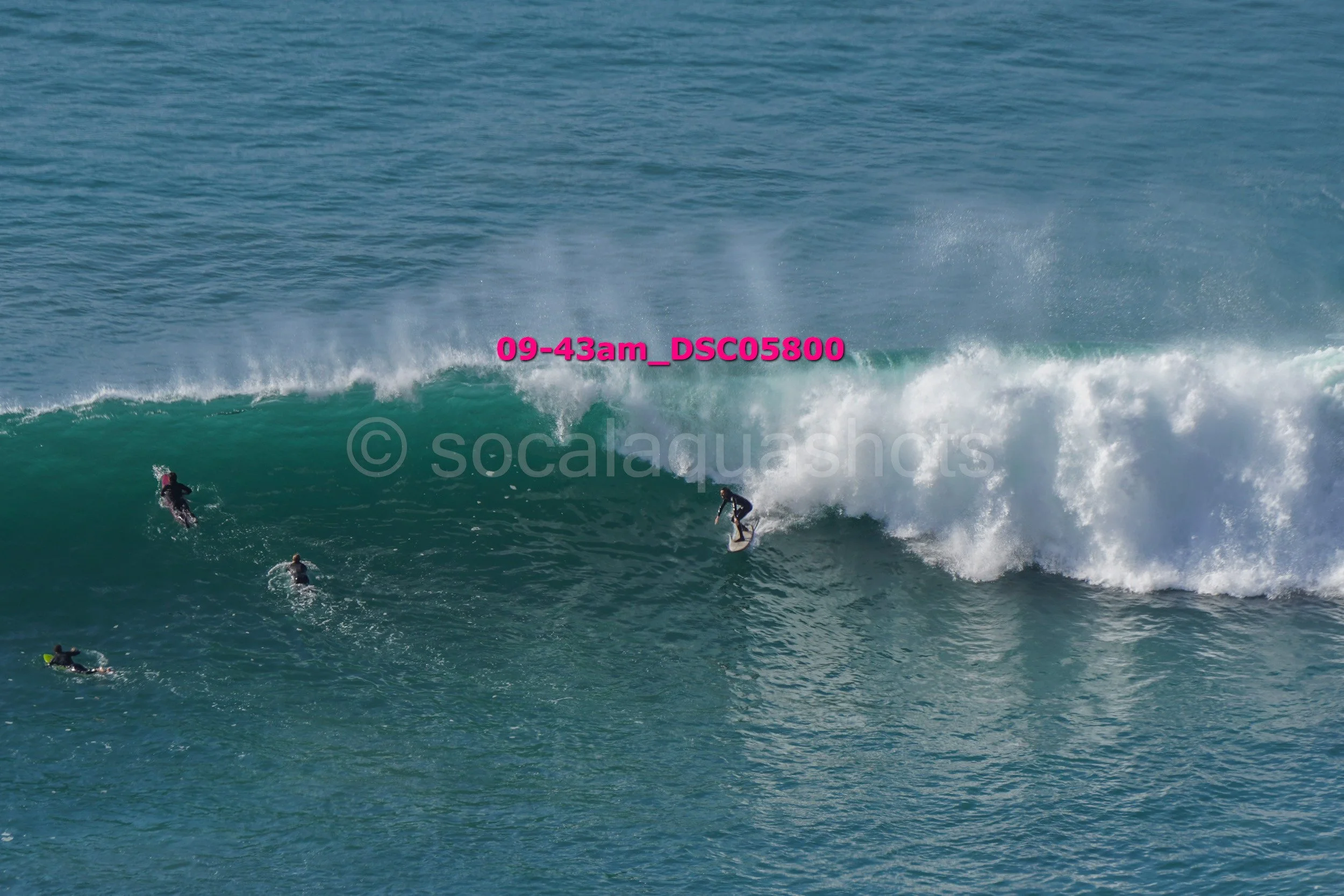 Surfers riding a large wave in the ocean, with one surfer standing on a surfboard on the crest of the wave and others paddling nearby.