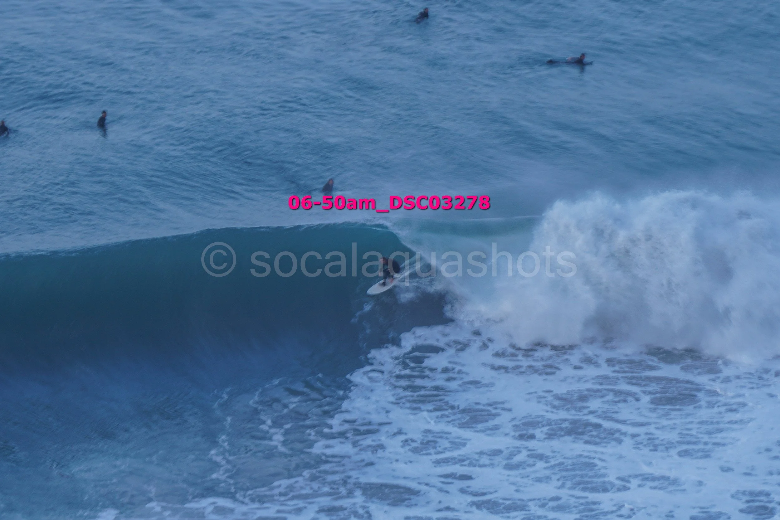 Surfer riding a large wave in the ocean, with several people swimming or surfing in the background.