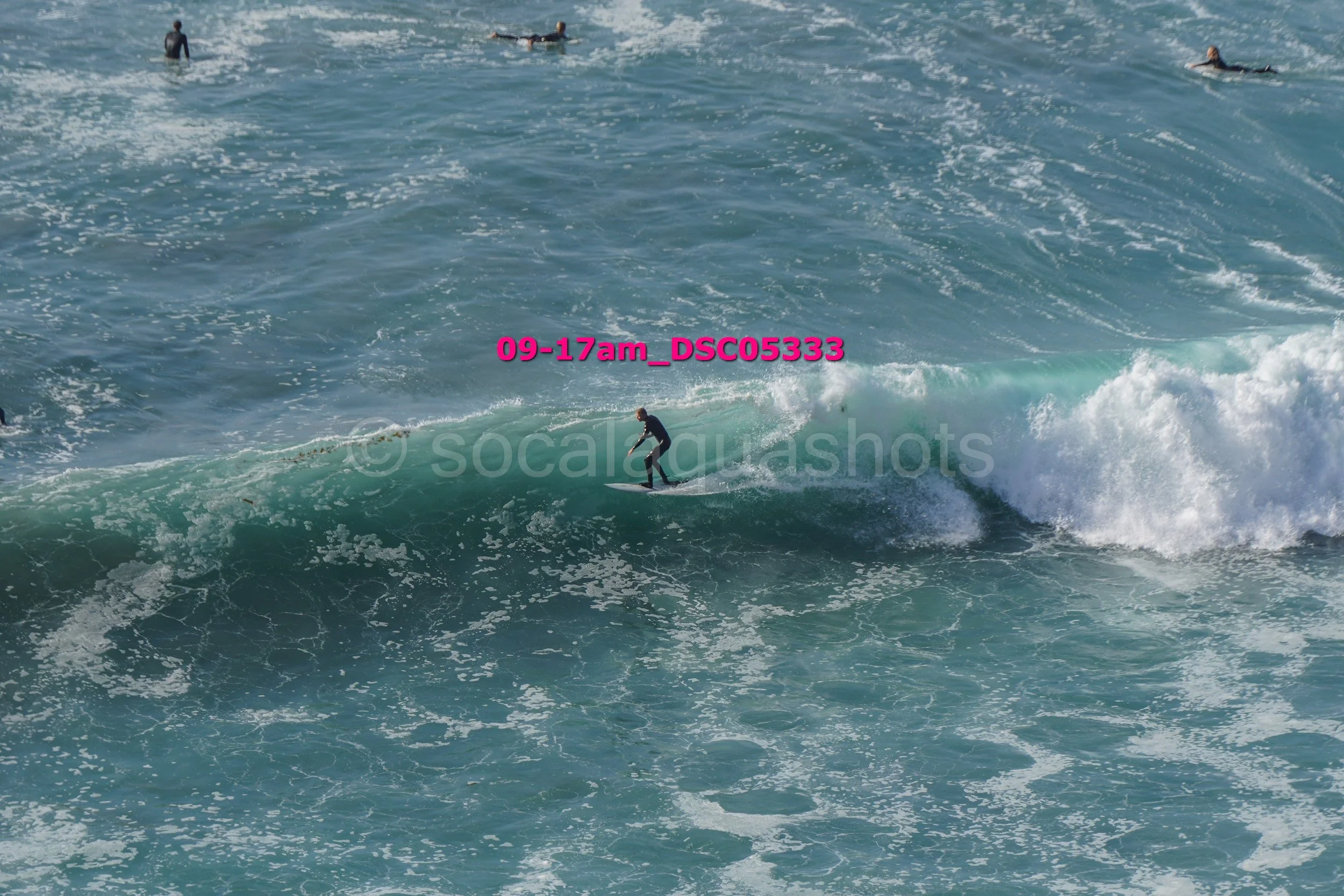 A person surfing on a large wave in the ocean with several other surfers visible in the background.