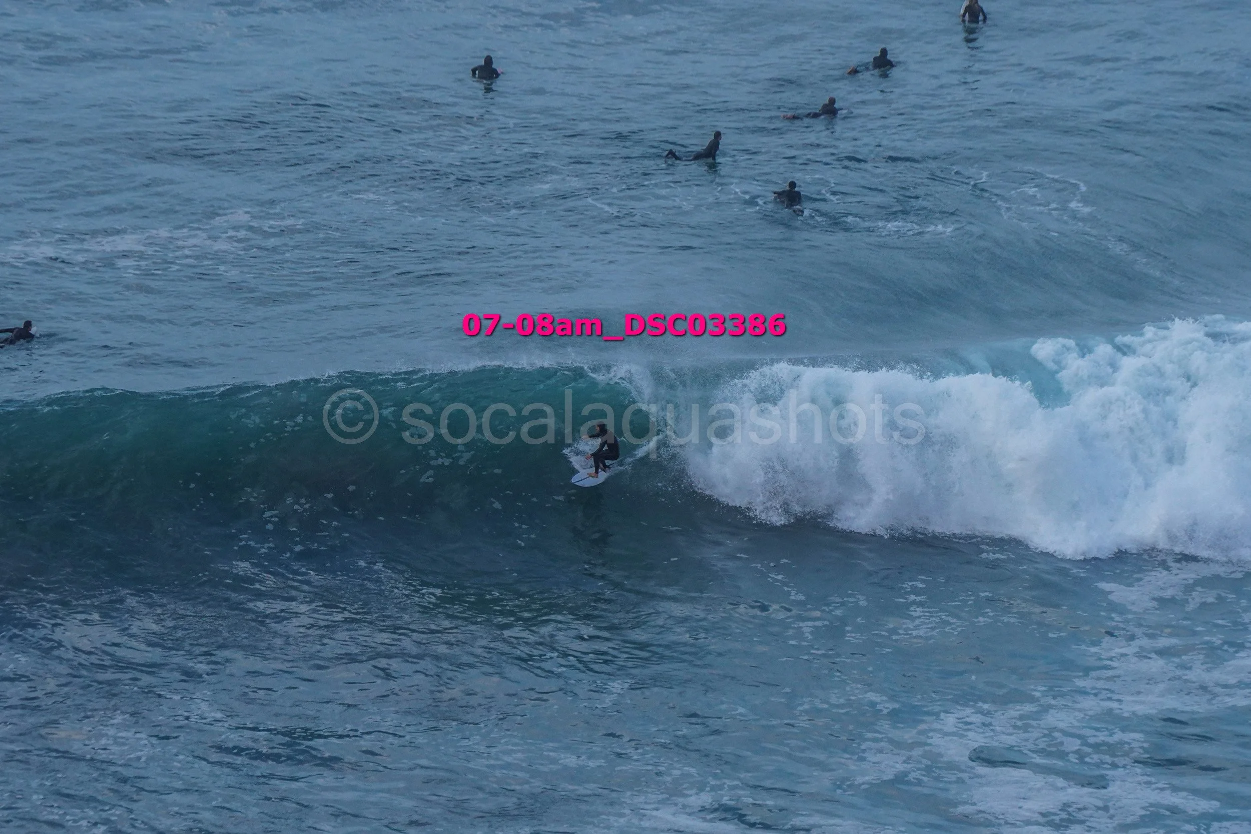 A surfer riding a wave in the ocean with multiple surfers and swimmers in the background.