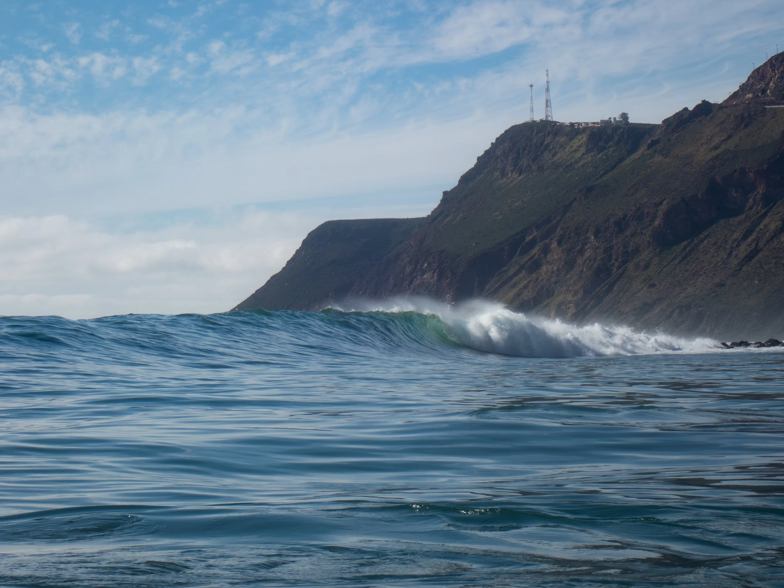 Ocean waves crashing near a rocky shoreline with cliffs in the background and a partly cloudy sky.