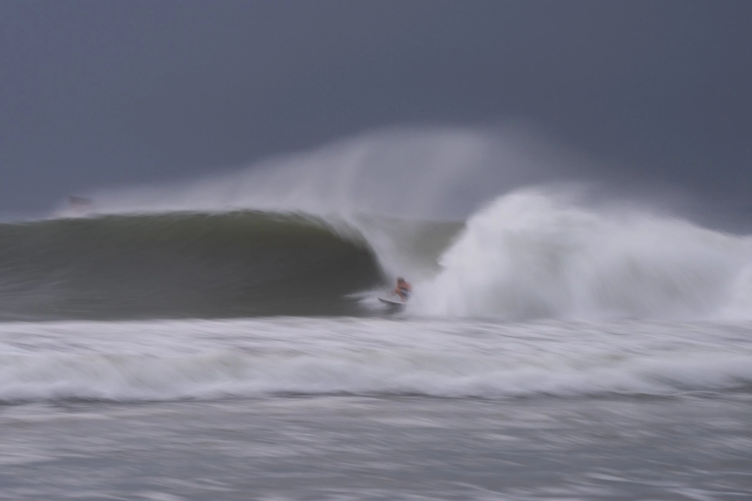 A surfer riding a wave with dark stormy clouds overhead, with a boat visible in the distance.