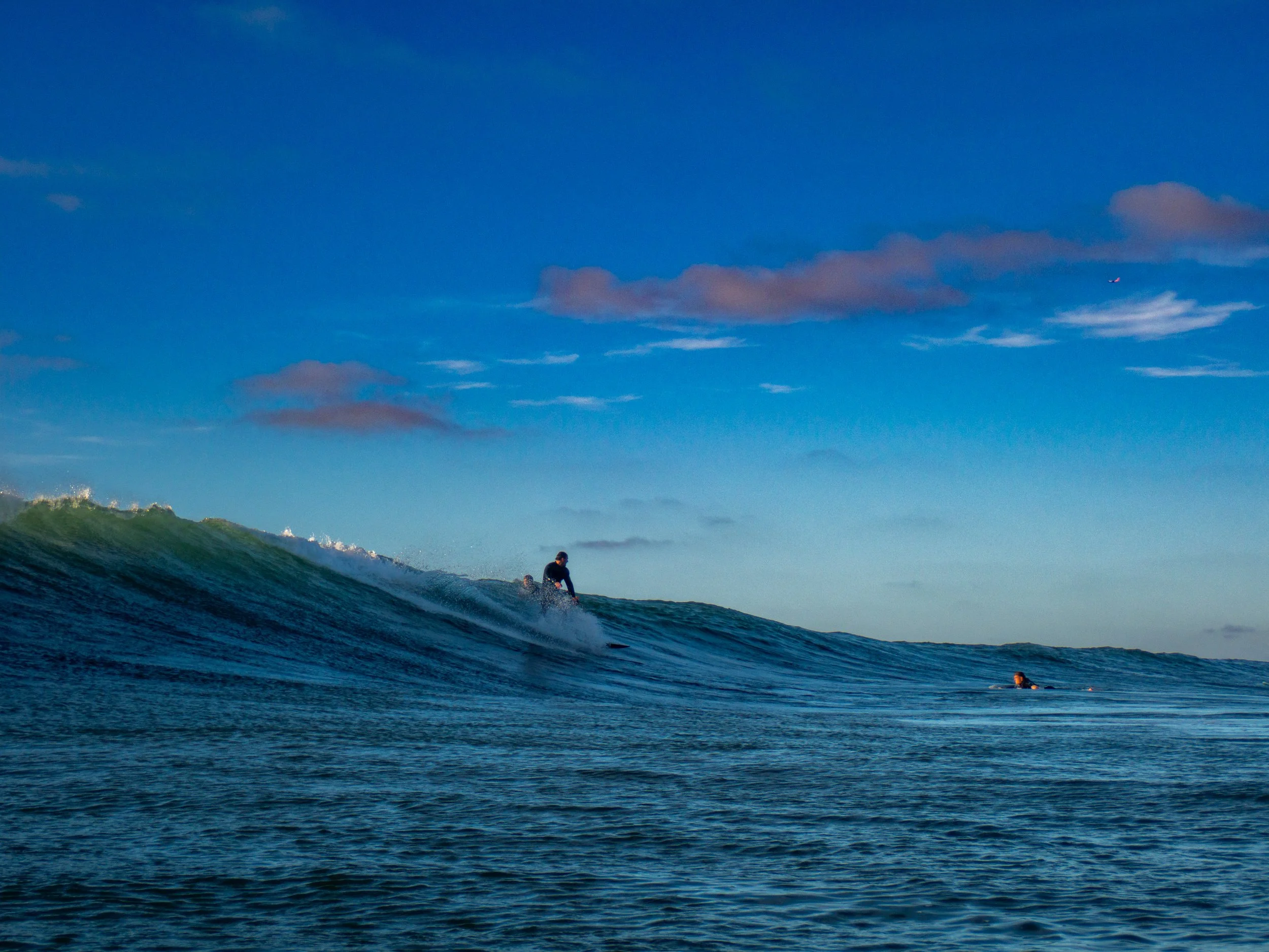A person surfing on an ocean wave with a clear blue sky and some clouds in the background.