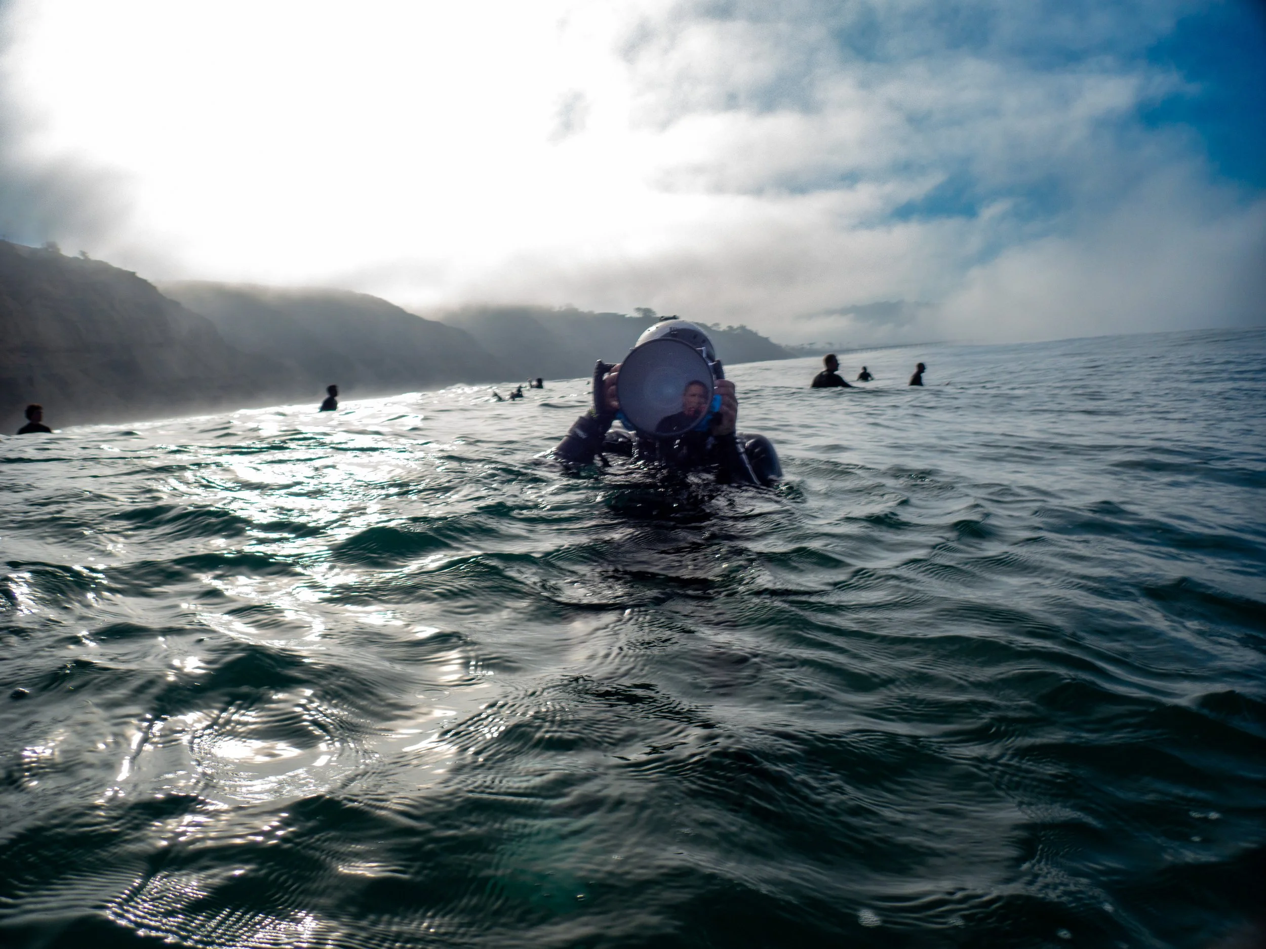 Person in wetsuit holding a mirror in the ocean with several other people swimming nearby and a cloudy sky in the background.
