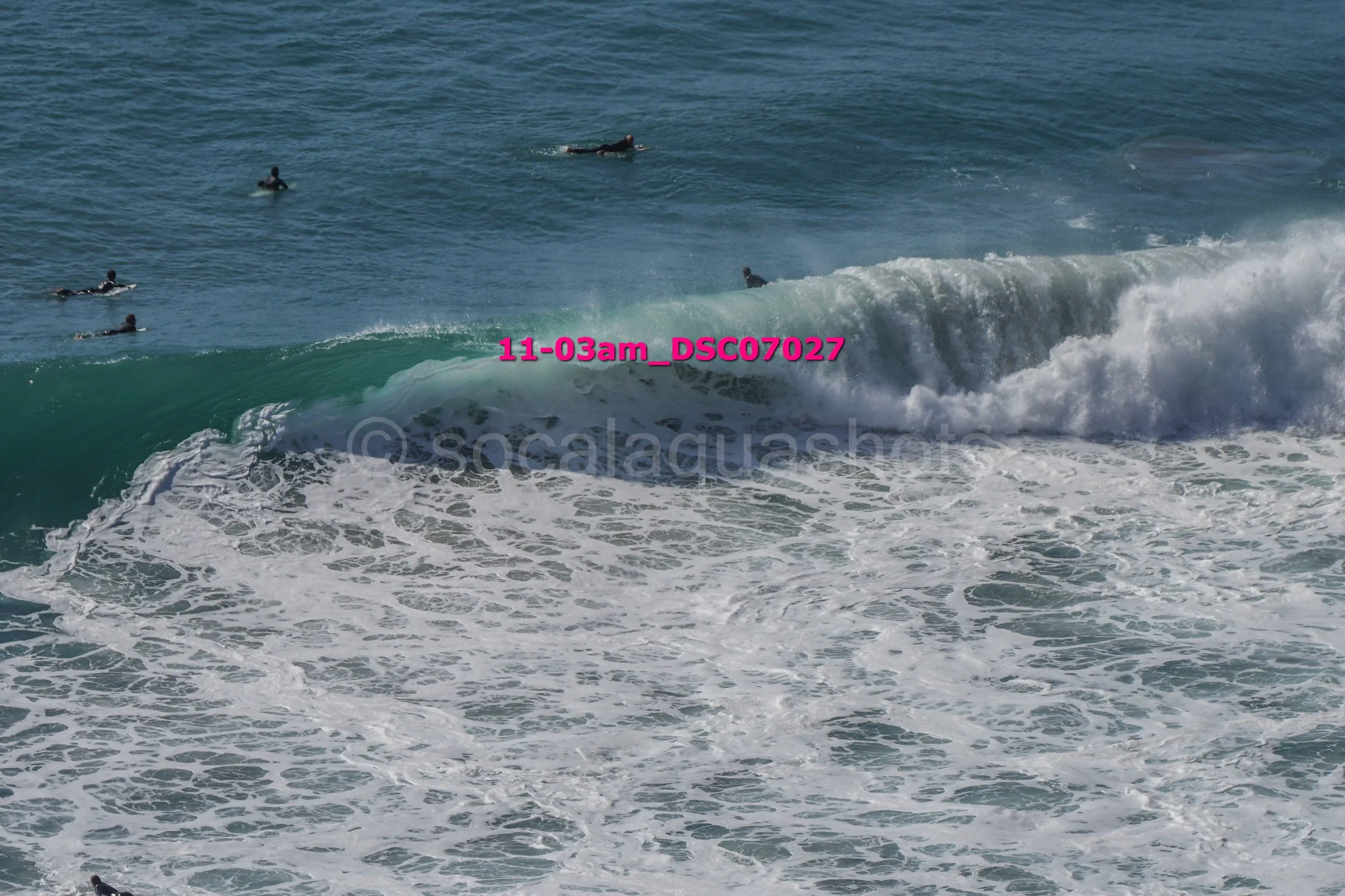 Surfers in the ocean, with one riding a wave and others waiting in the water, amid foamy surf and blue sea.
