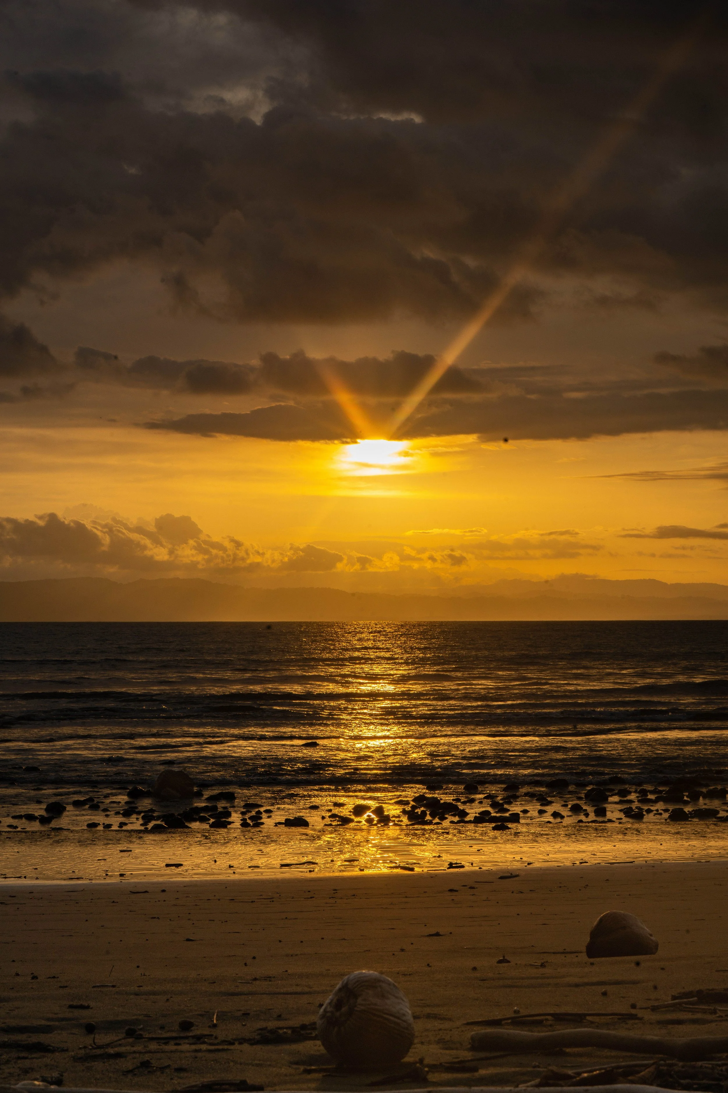 Sunset over the ocean with clouds in the sky, reflecting on the water, and large rocks on the sandy beach in the foreground.