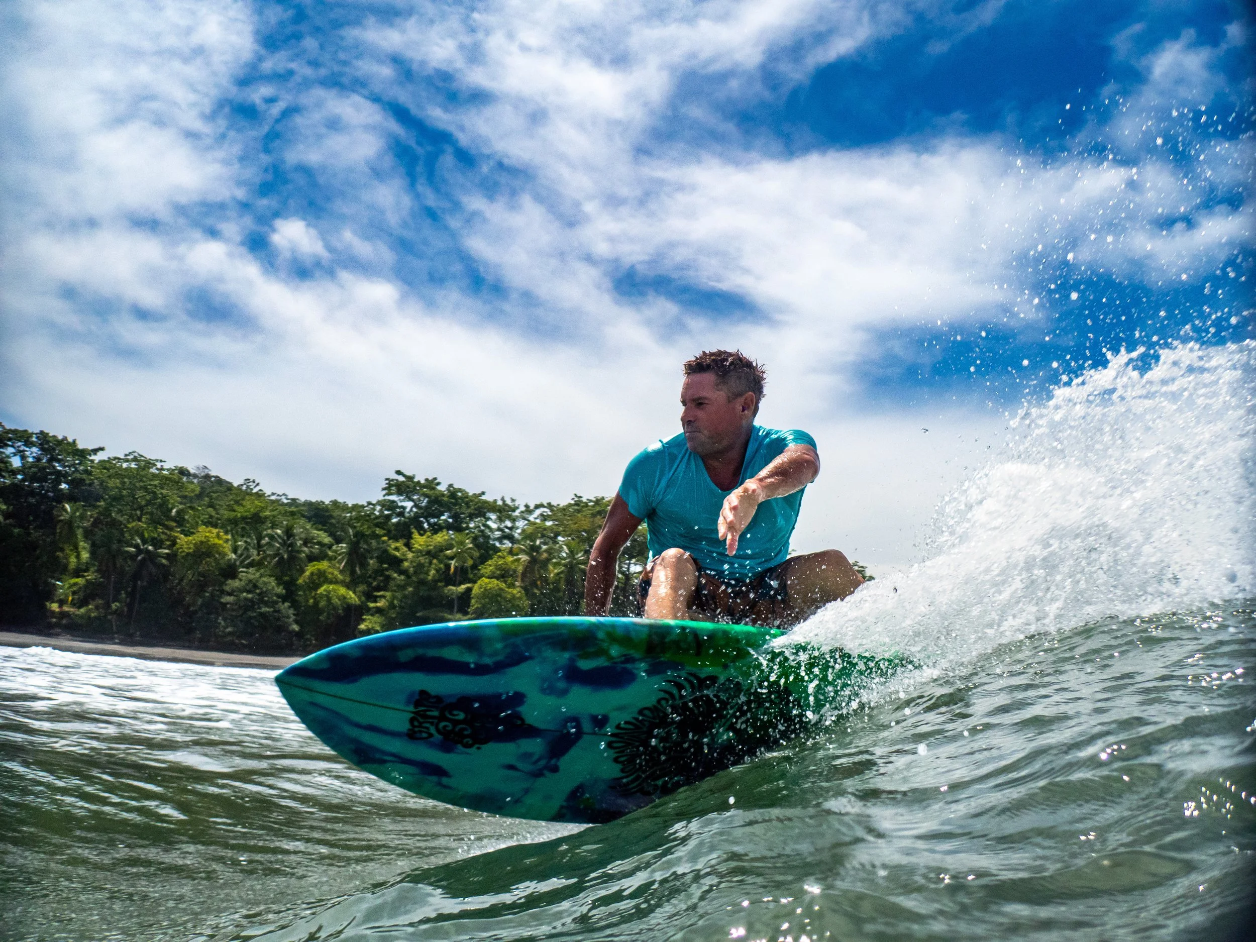 Man surfing on a wave in the ocean with a tropical forest and partly cloudy sky in the background.