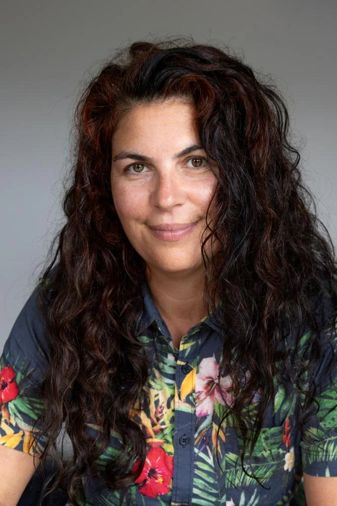 Person with long curly hair wearing a floral patterned shirt, smiling slightly against a plain background.
