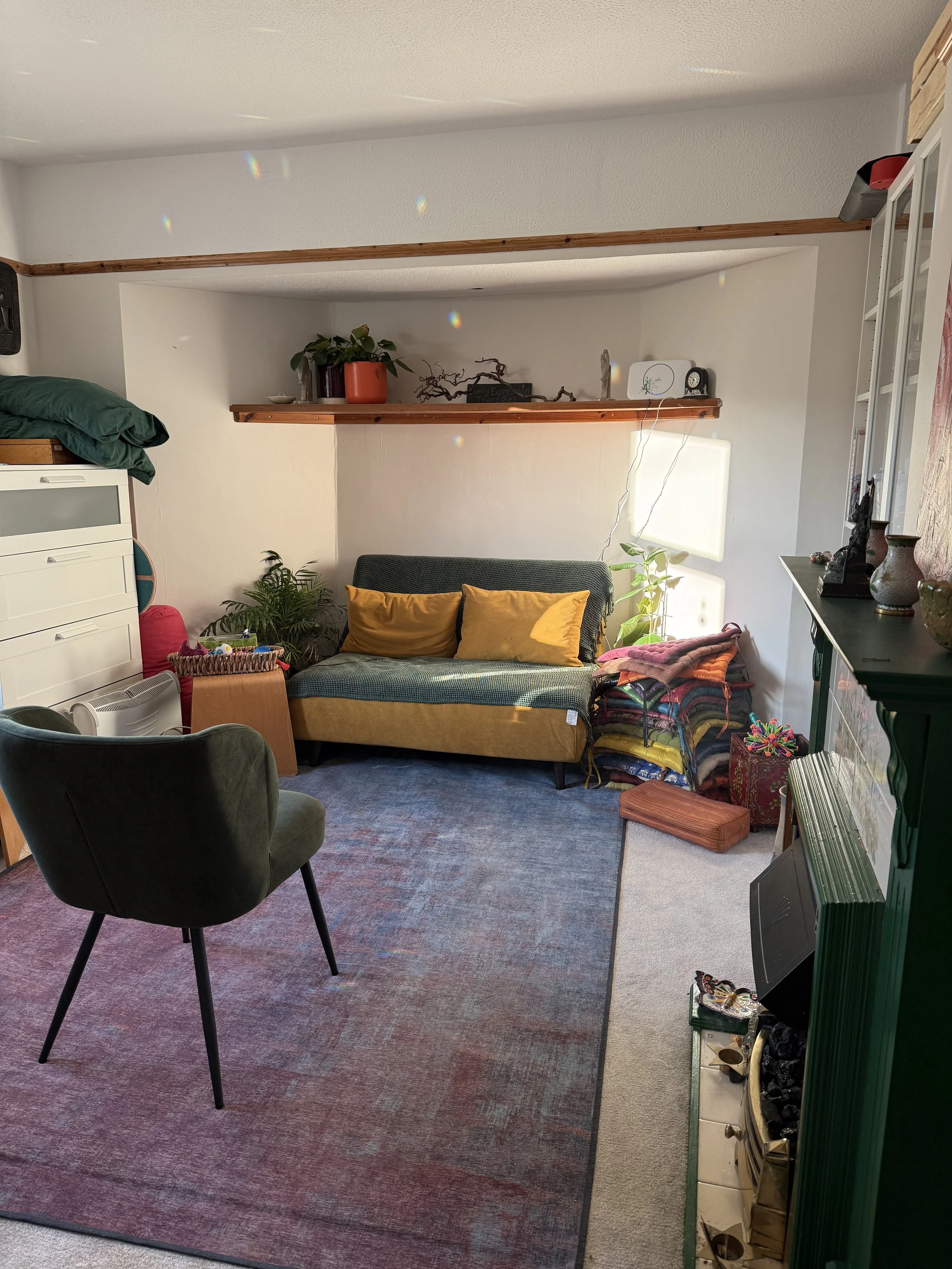 The therapy space at Every Expression Therapies. A dark green chair sits in front of a small yellow sofa. There is a fireplace in the room and lots of natural light.