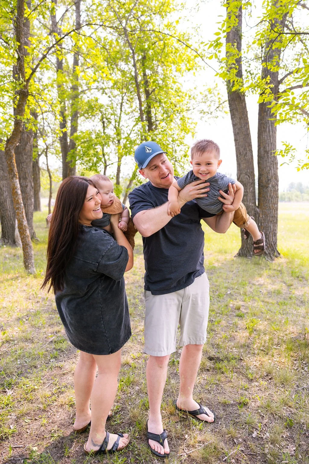 A family of four enjoying a sunny day in a forest, with the father lifting a baby girl, the mother and older daughter sitting on the grass looking at the baby.