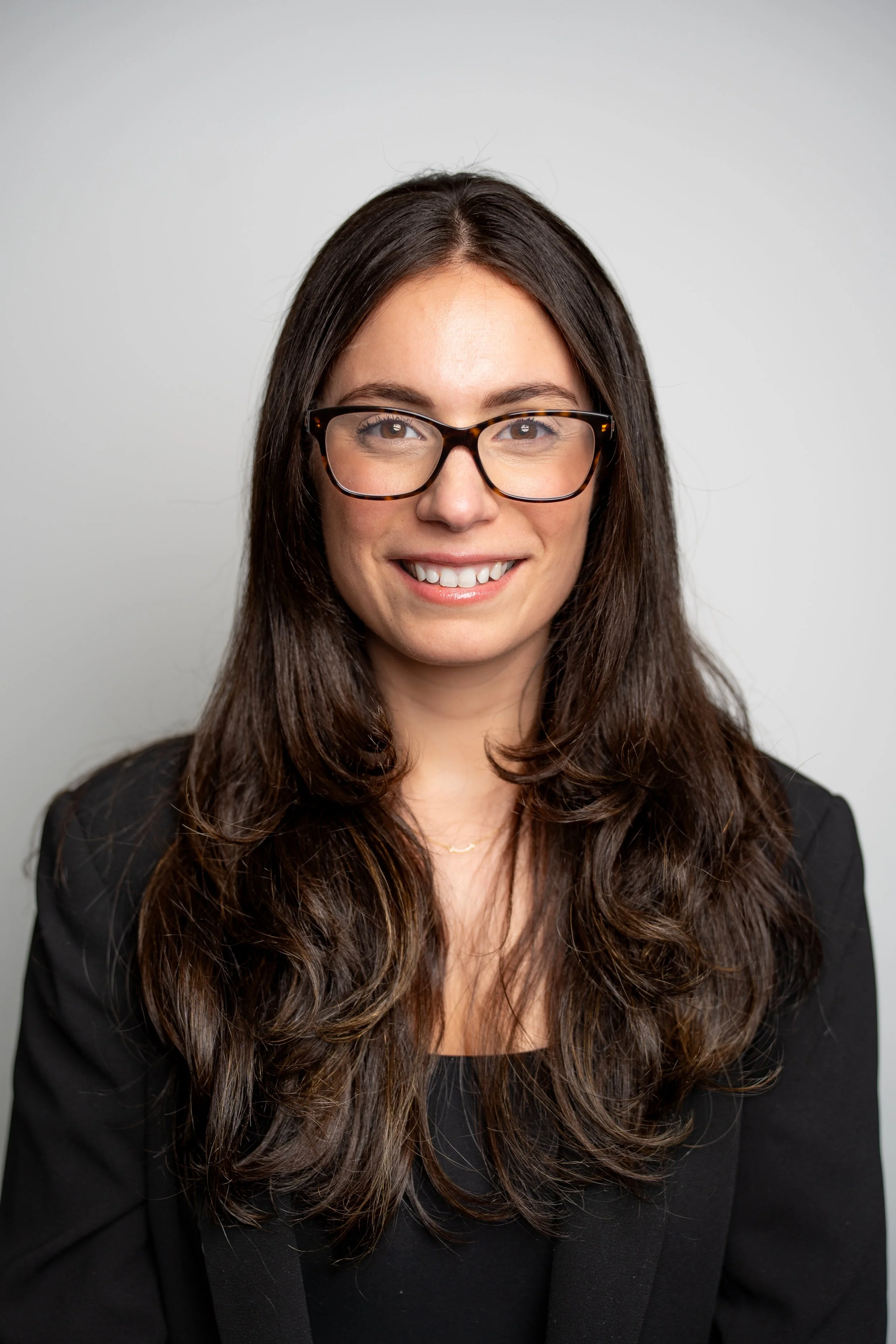 A woman with long, wavy brown hair, wearing glasses and a black blazer, smiling at the camera against a neutral gray background.