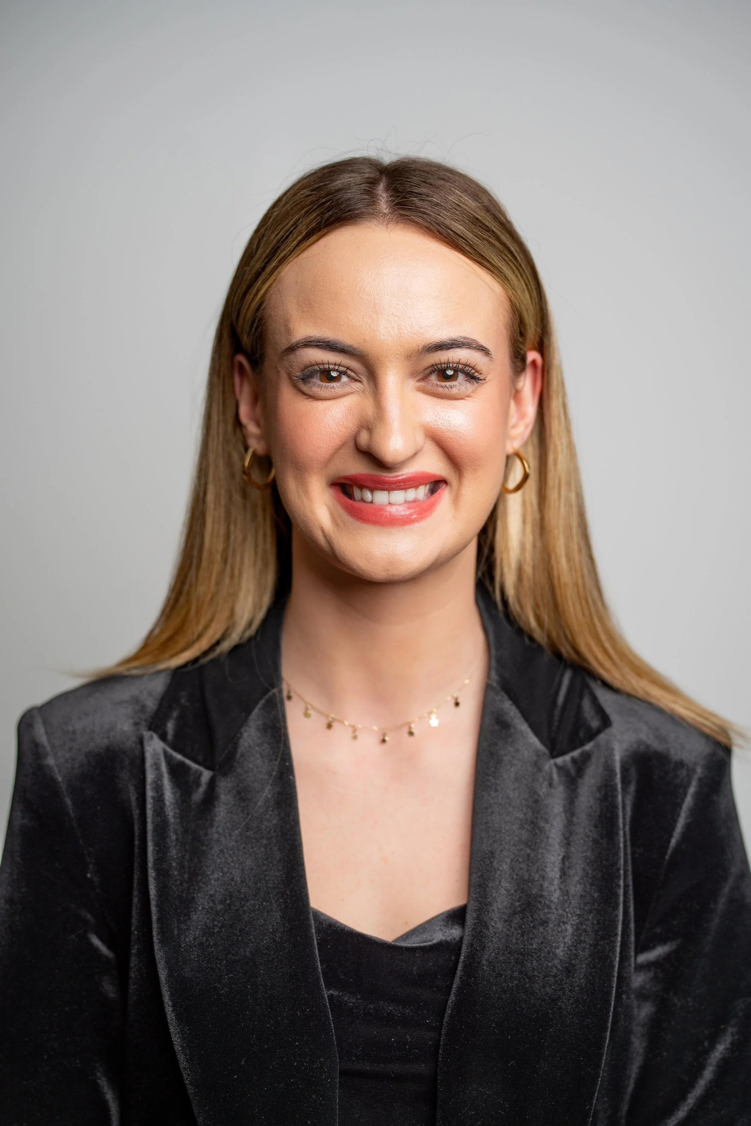 A woman with blonde hair smiling, wearing gold hoop earrings, a necklace, and a dark velvet blazer.