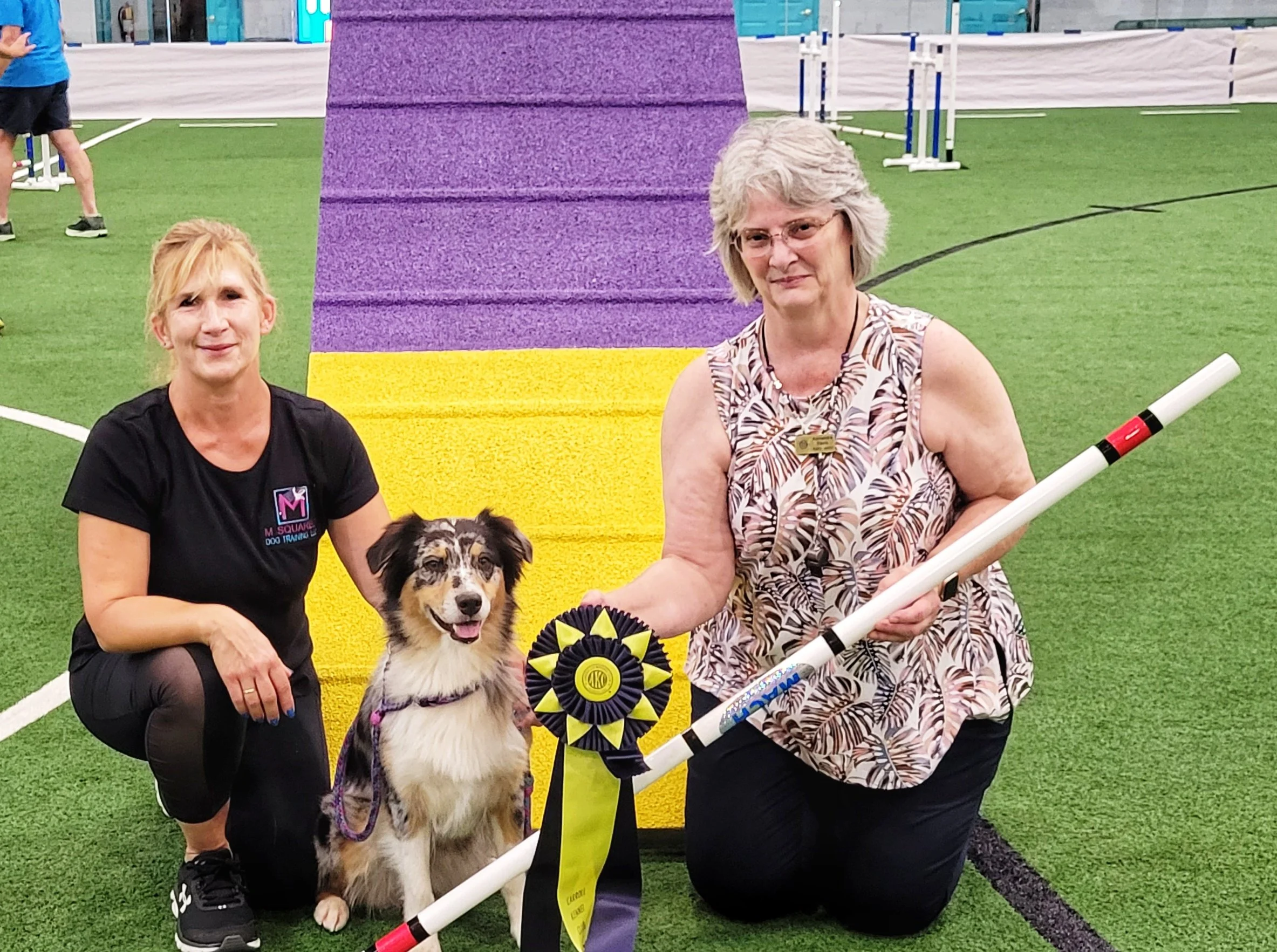 Two women with an Australian Shepherd dog sitting between them on an indoor field.