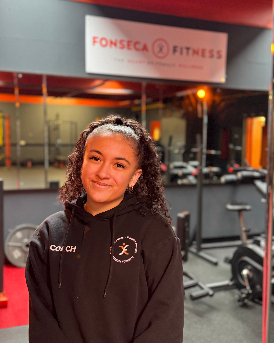 A young woman with curly hair tied back in a ponytail, smiling at the camera, in a gym setting. She is wearing a black hoodie with 'COACH' printed on it and the FONSECA FITNESS logo. Gym equipment, mirrors, and a sign reading 'FONSECA FITNESS' are visible in the background.