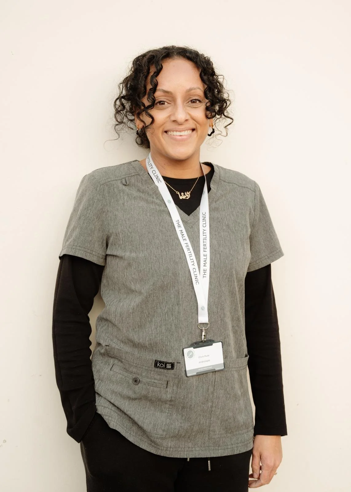 A woman with curly hair wearing a gray medical scrub top, black long-sleeve shirt, a name tag, and a lanyard that says 'The Male Fertility Clinic.'