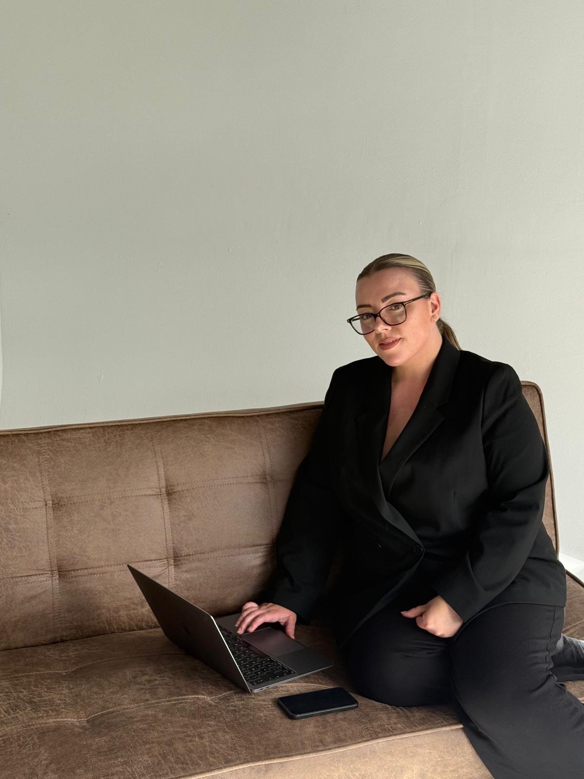 A woman wearing glasses and a black blazer, sitting on a tan couch with a laptop and smartphone in front of her, against a plain light-colored wall.