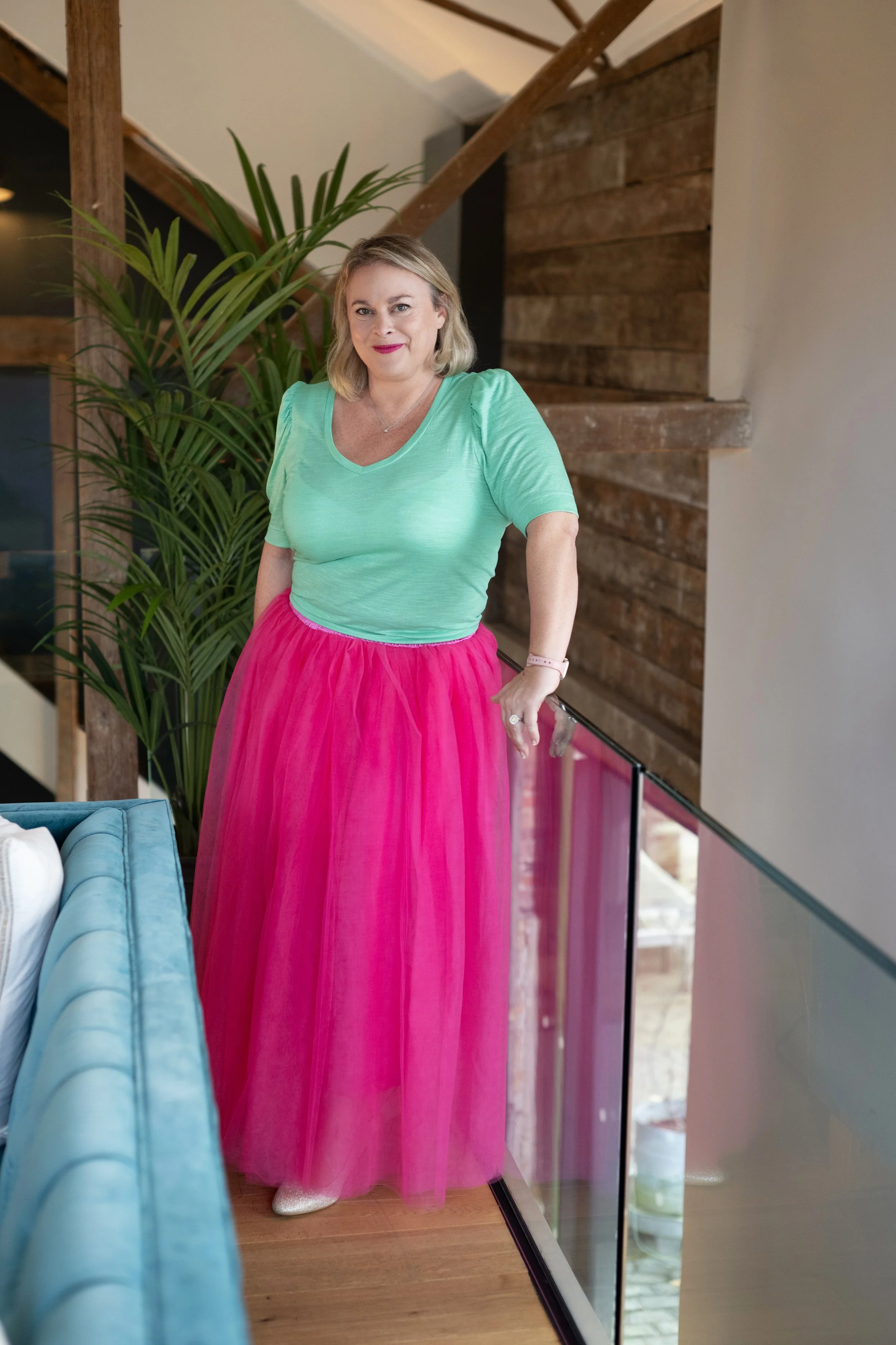 A woman standing on a balcony next to a large green plant, wearing a mint green top and a pink tulle skirt, indoors with wooden wall accents.