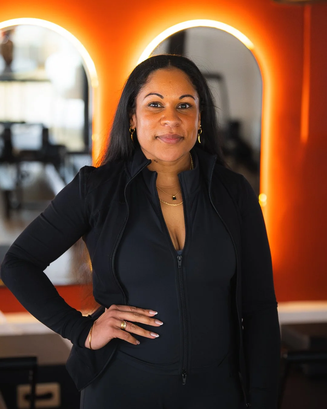 A woman with medium skin tone and long black hair wearing a black athletic jacket and gold jewelry stands in front of illuminated mirror in a modern interior.