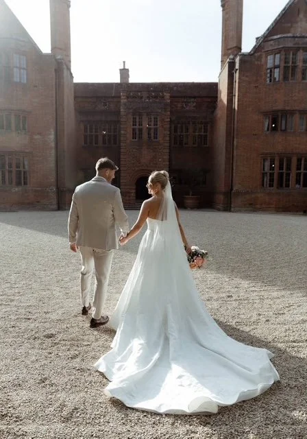 A bride in a white wedding gown and a groom in a light-colored suit holding hands in a courtyard of a historic brick building.