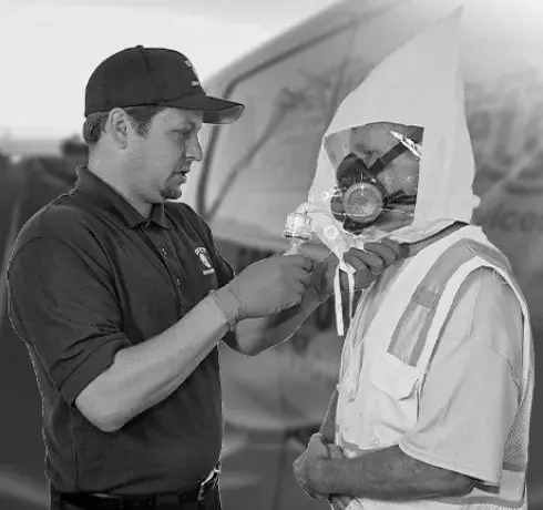 A man in a black cap adjusting a person's protective helmet with a face shield, both standing outdoors near a vehicle.