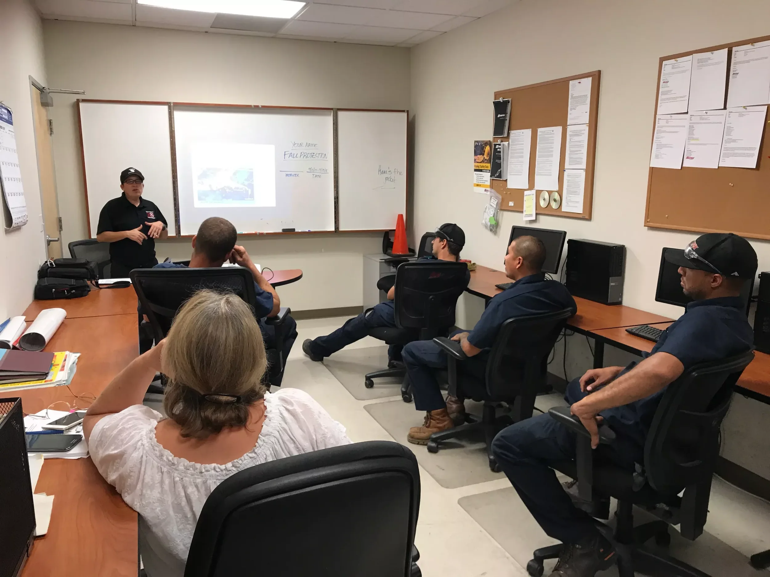 A group of five people in a classroom watching a presentation at the whiteboard, with one woman sitting at a desk in the foreground, and four men sitting in chairs facing the front.