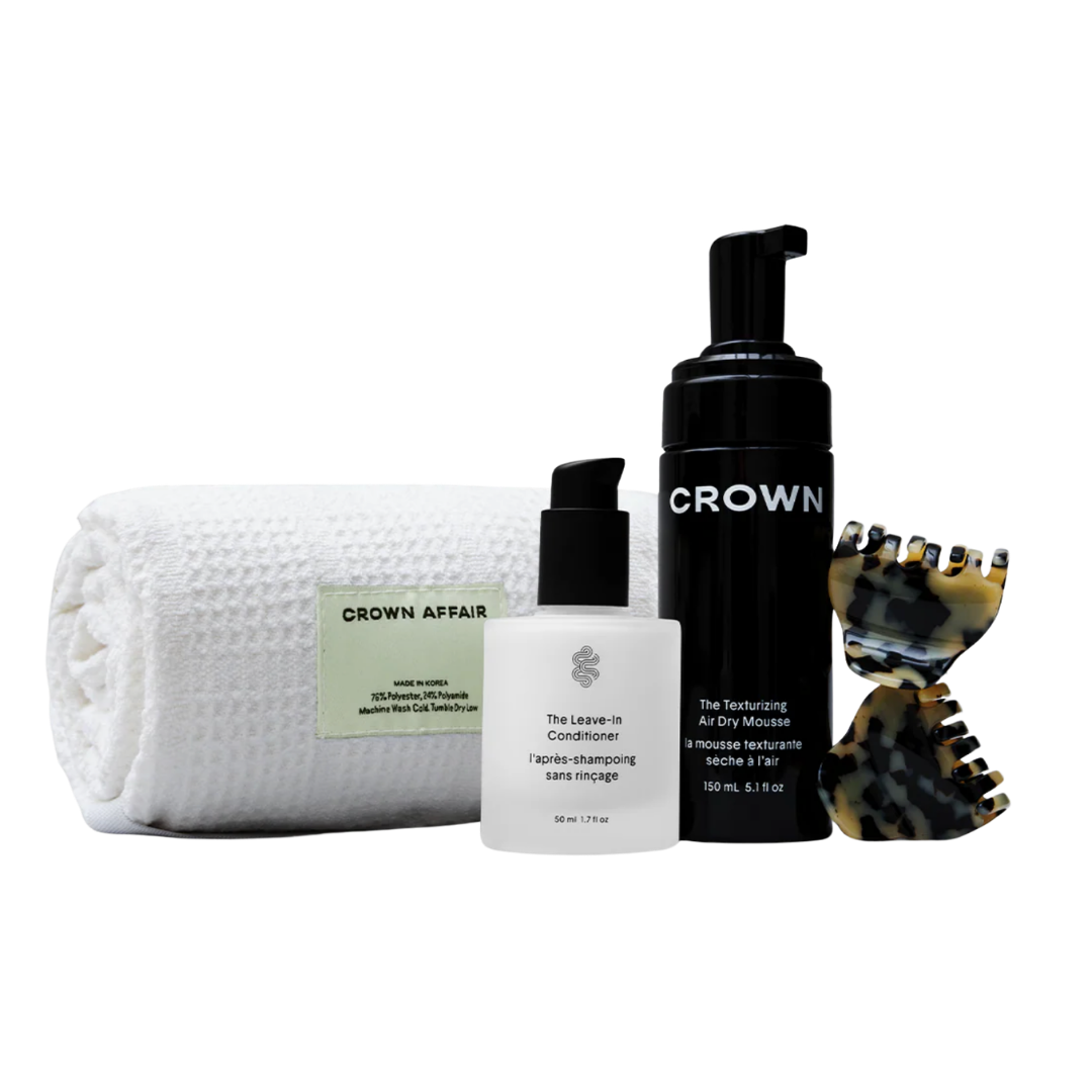 A Crown Affair hair care set including a white waffle-knit towel, a white pump bottle, a black bottle of shampoo, and a tortoiseshell hair clip.