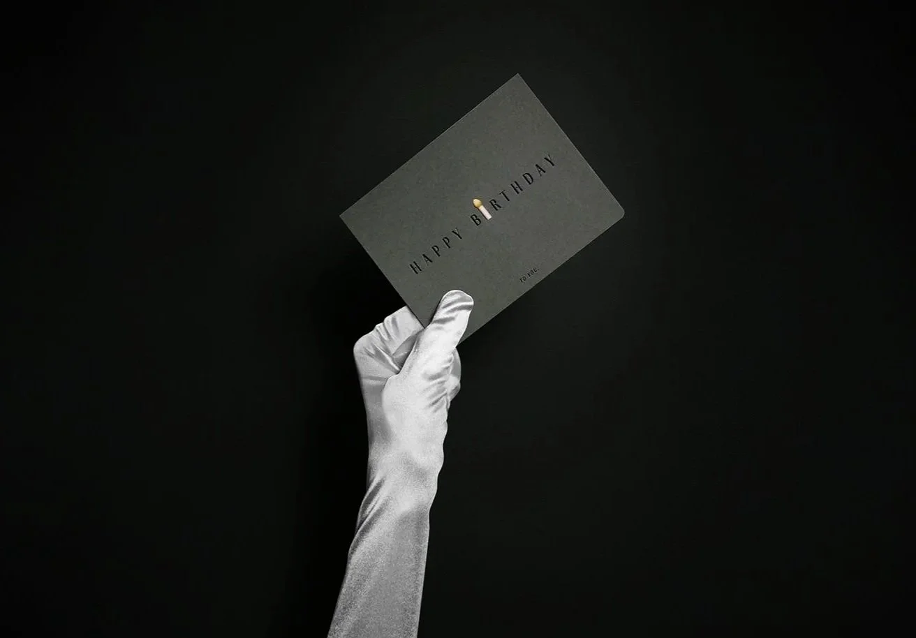 A hand wearing a long silver satin glove holds up a dark grey 'Happy Birthday' card against a black background.