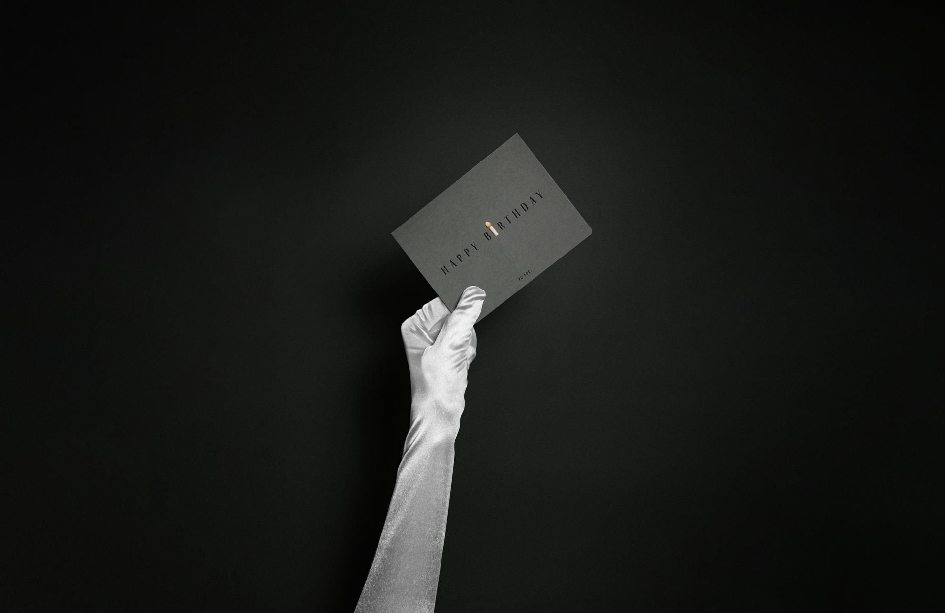 A hand wearing a long silver satin glove holds up a dark grey 'Happy Birthday' card against a black background.