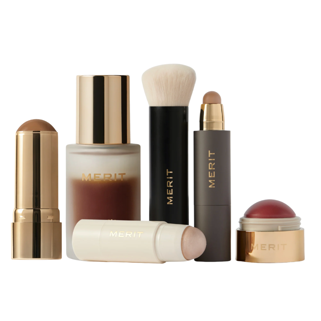A collection of Merit Beauty products including a foundation stick, a bronze stick, a blending brush, and a small gold jar of lip/cheek tint.