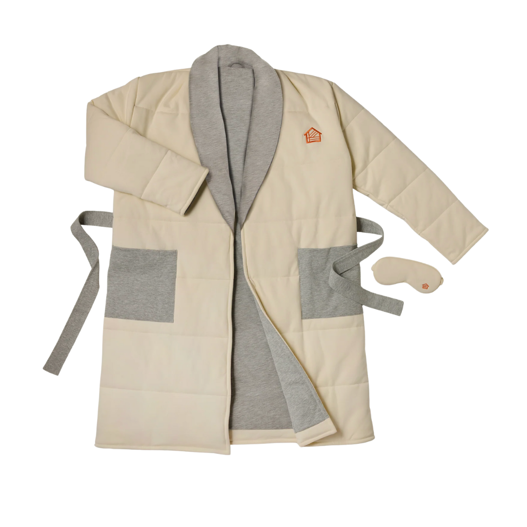 A quilted, cream-colored luxury housecoat with grey pocket accents, shown with a matching padded sleep mask.