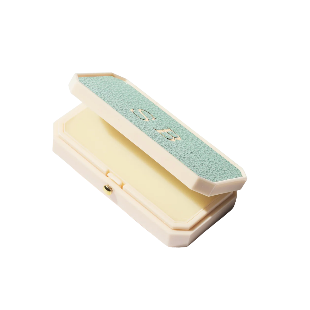 An open cream-colored lip balm compact with a textured light blue lid featuring gold initials.