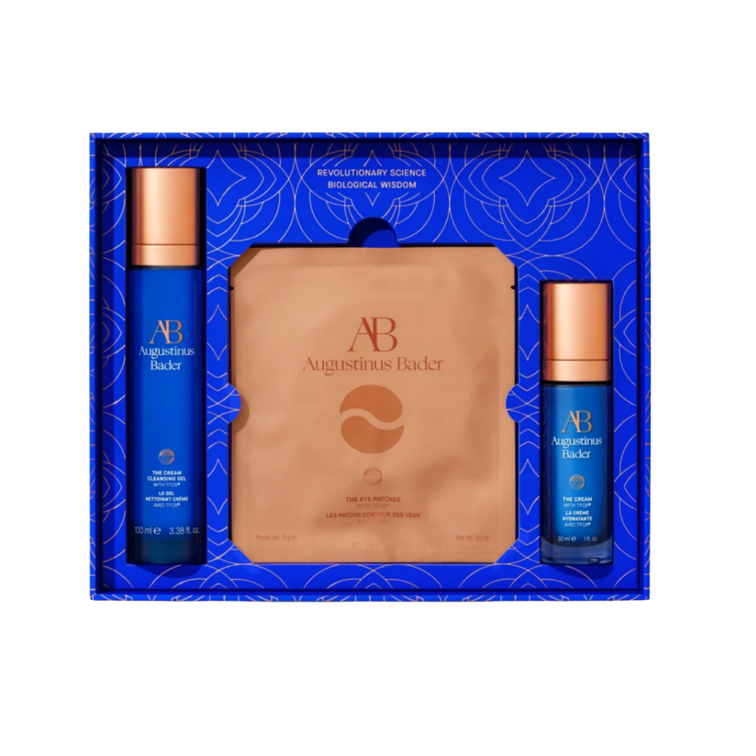 An Augustinus Bader skincare set in a vibrant blue and gold box, featuring a full-size serum, eye cream, and copper-toned eye patches.
