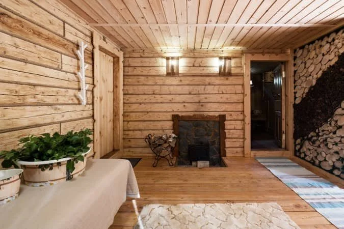 Rustic wooden sauna interior with log walls, wooden ceiling and floor, a small stone fireplace, a table with white cloth and potted plants, and a wall with stacked firewood.