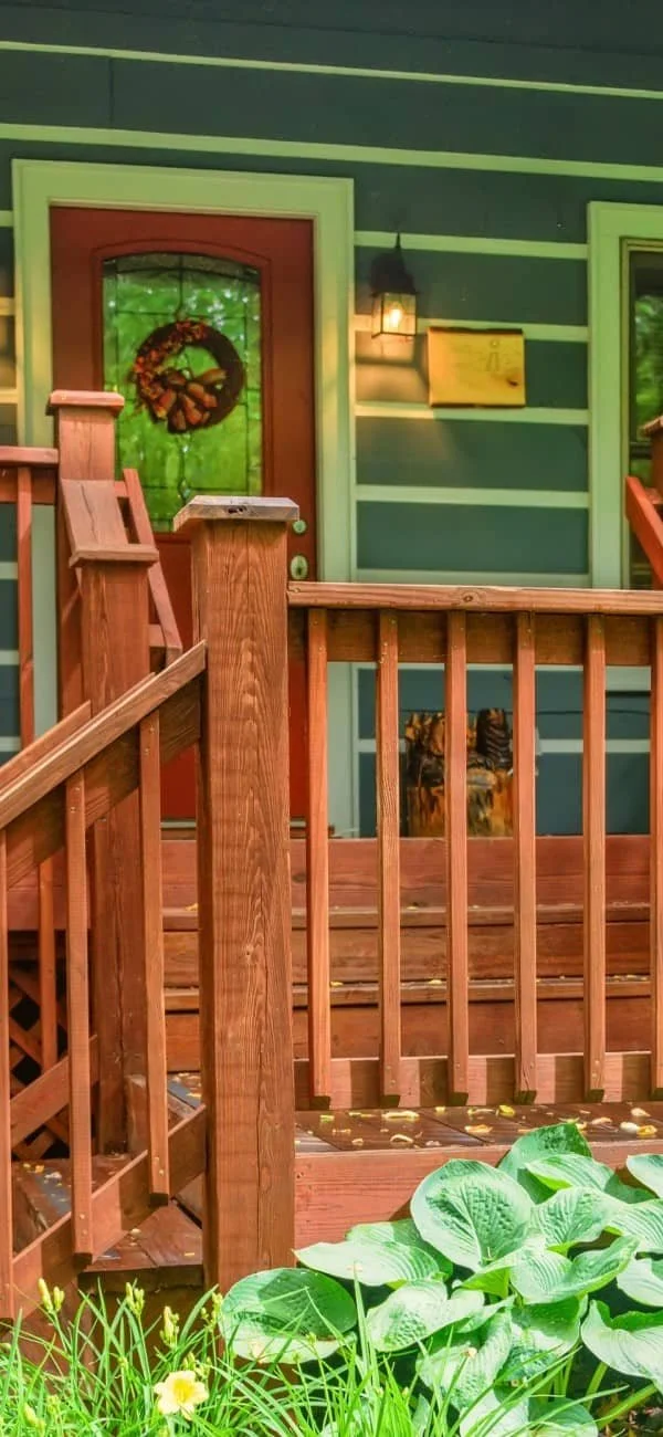 Wooden porch with railing, front door with wreath, and green plants in foreground.