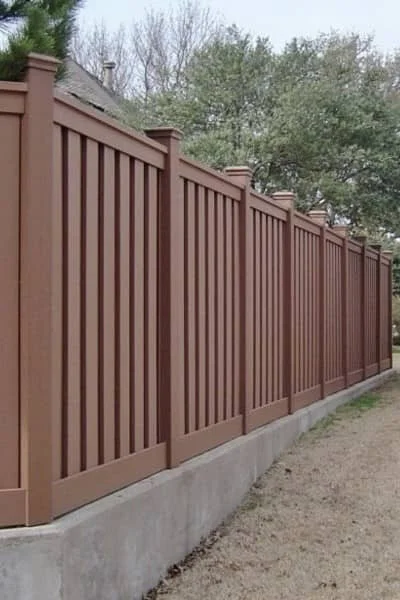 Brown vinyl fence with vertical slats on a concrete base, set against trees and an overcast sky.