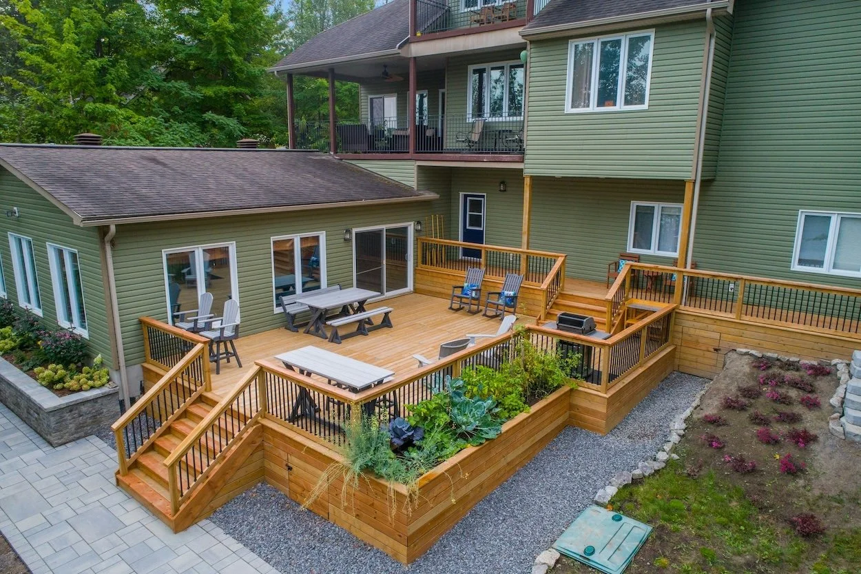 Vista aérea de una casa con una Terraza madera con muebles de exterior, camas de jardín y una barbacoa.