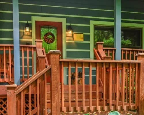 Wooden front porch with railing and steps, light green siding, door with wreath, two windows, and decorative lights on either side of the door.