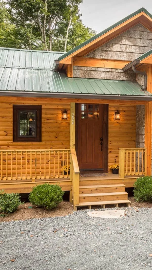 Front view of a rustic log cabin with a green metal roof, wooden porch, and steps. Two outdoor lanterns flank the entrance, and potted plants decorate the porch area. There's a gravel pathway leading to the cabin, surrounded by bushes and trees.