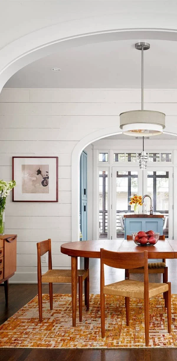 Dining room with a wooden table, four chairs, and a modern pendant light. A bowl of red apples is on the table. Light-colored walls, an artwork, and an interior archway leading to a kitchen area are visible.