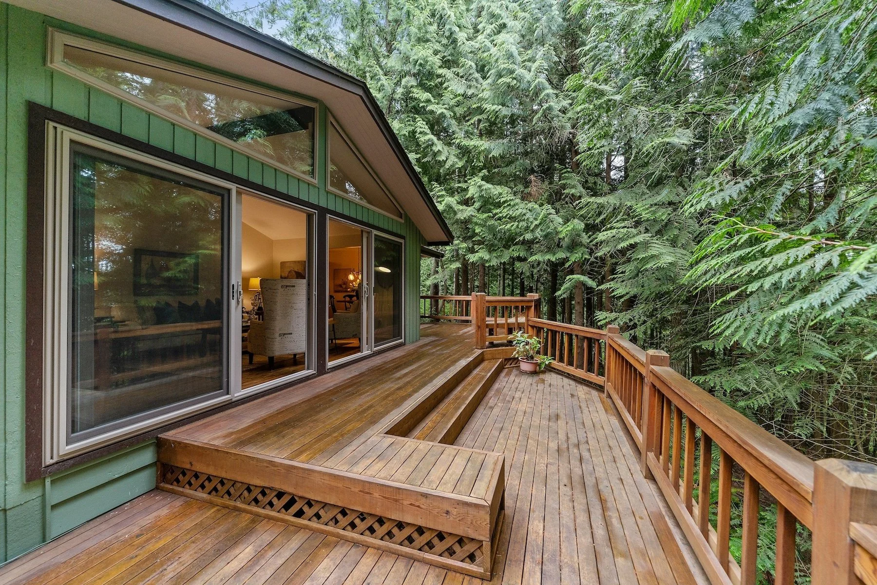 Terraza de madera con barandilla (barandal), rodeada de árboles, adosada a una casa con grandes puertas correderas de cristal.