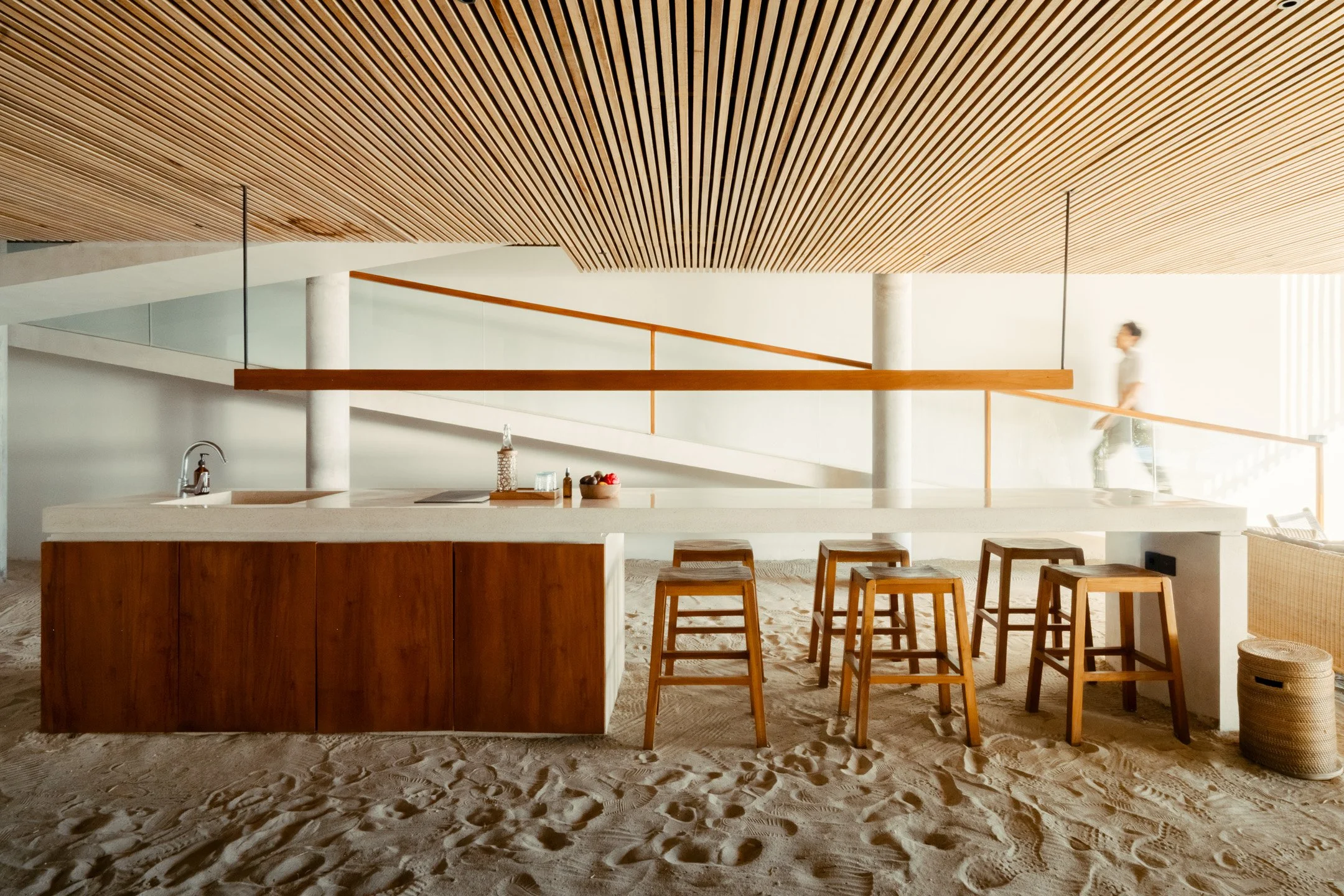 Interior of a modern beach house with a wooden ceiling, a kitchen island with stools, and a person walking past a glass railing on a sandy floor.