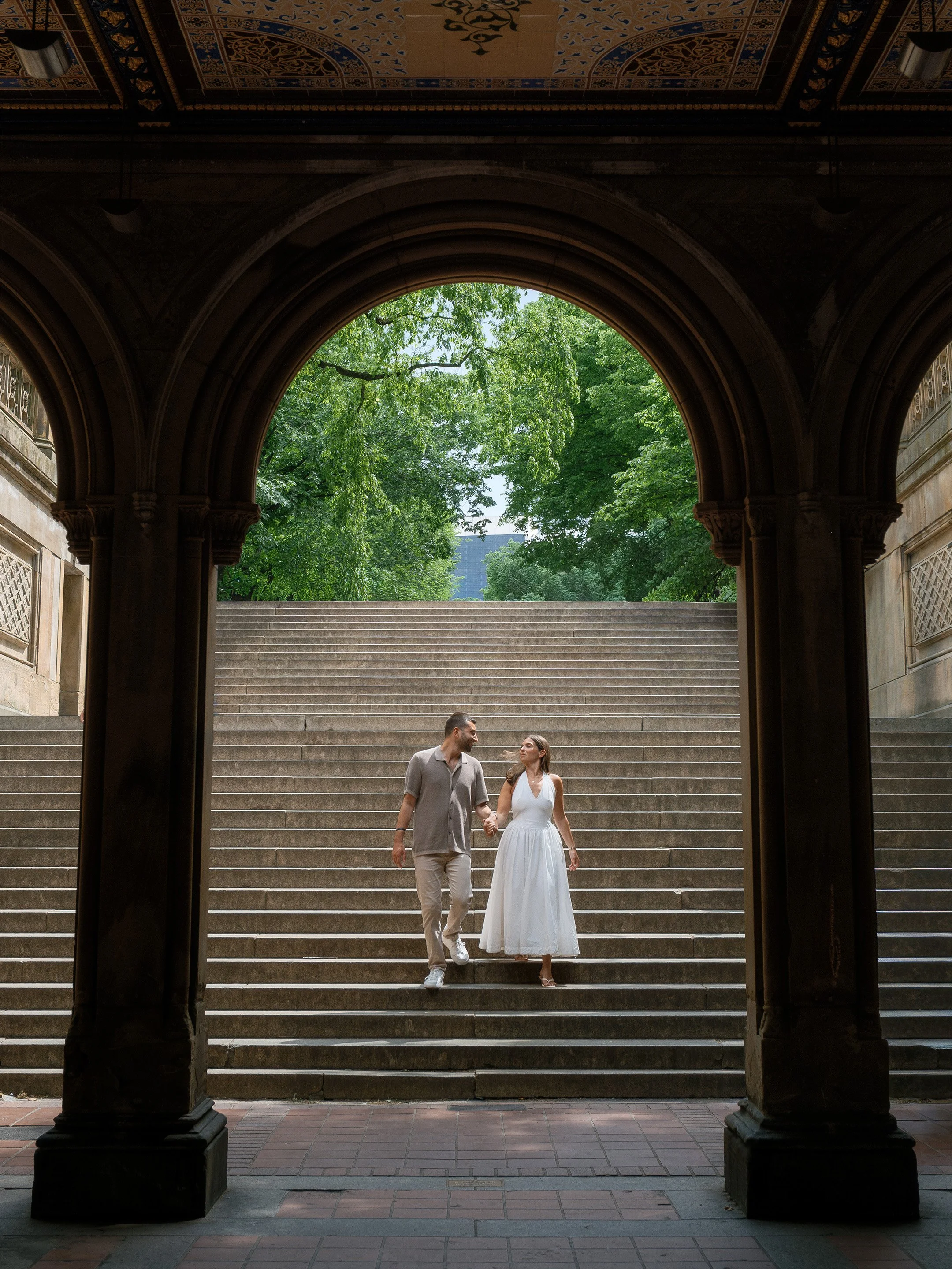 A couple walking down stairs outside, holding hands, with a large archway and lush green trees in the background.