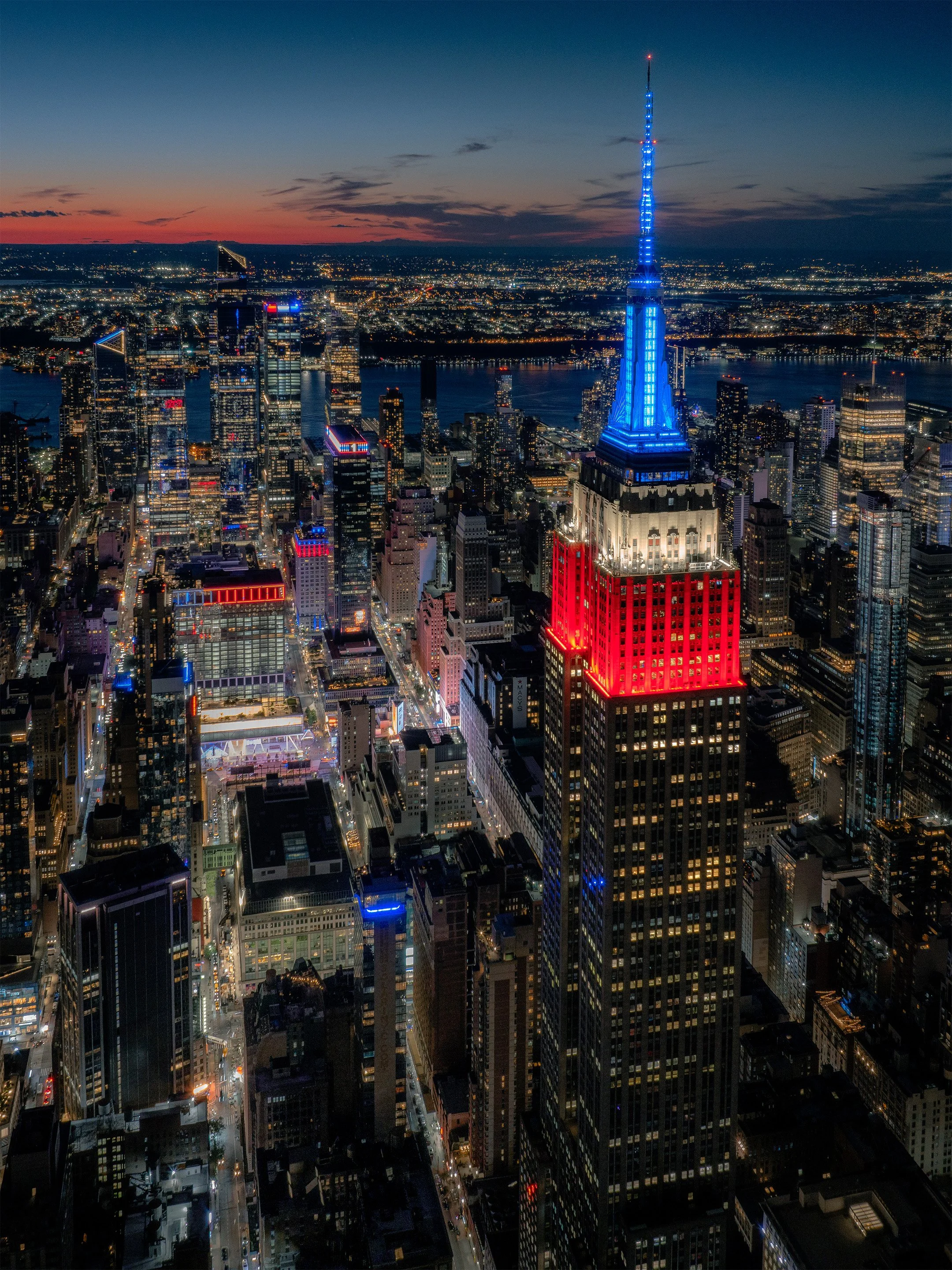 Nighttime cityscape of New York City with the Empire State Building lit up in red, white, and blue, and a skyline filled with illuminated skyscrapers.
