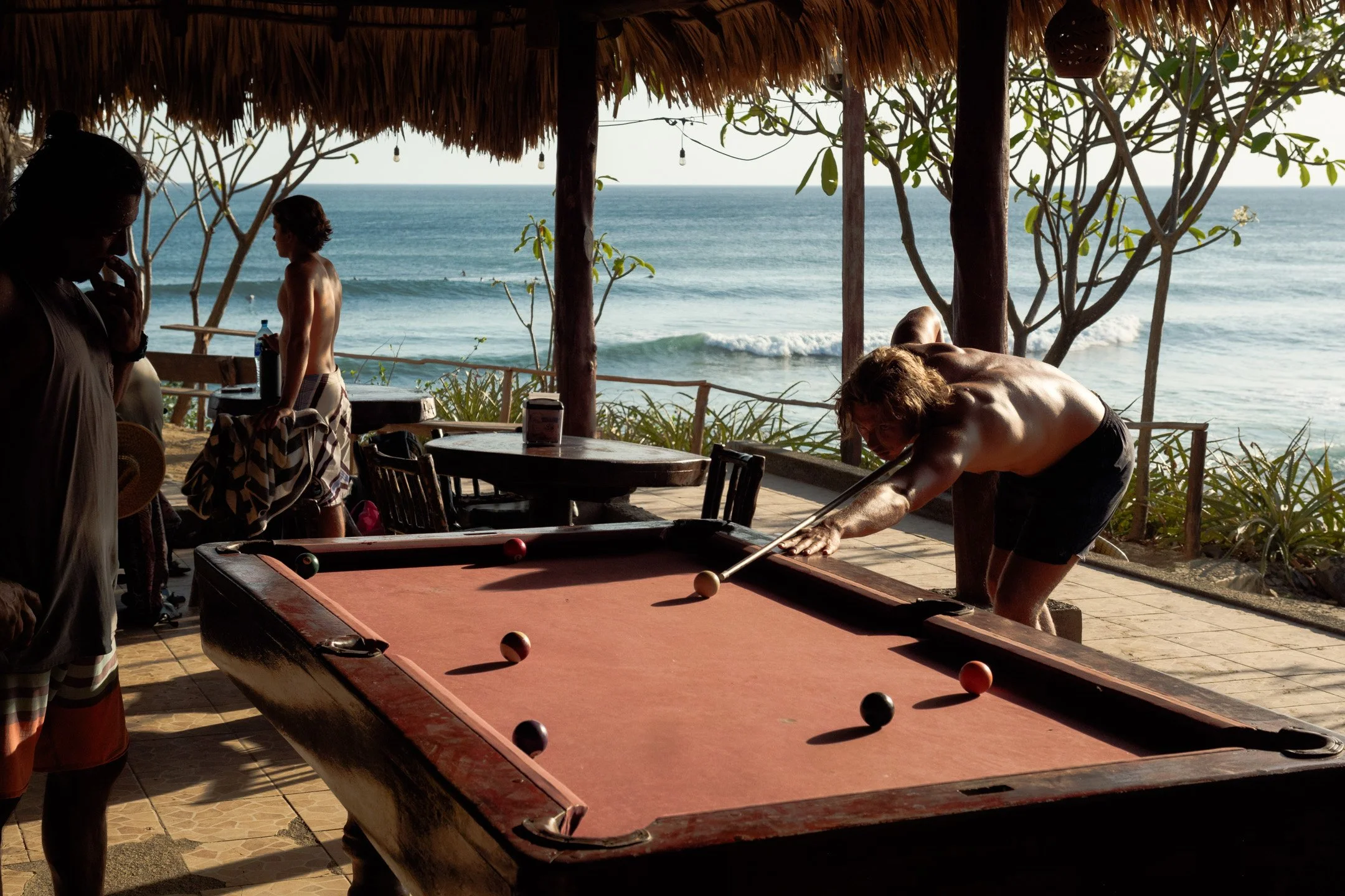 People playing pool under a thatched-roof hut with a view of the ocean in the background.