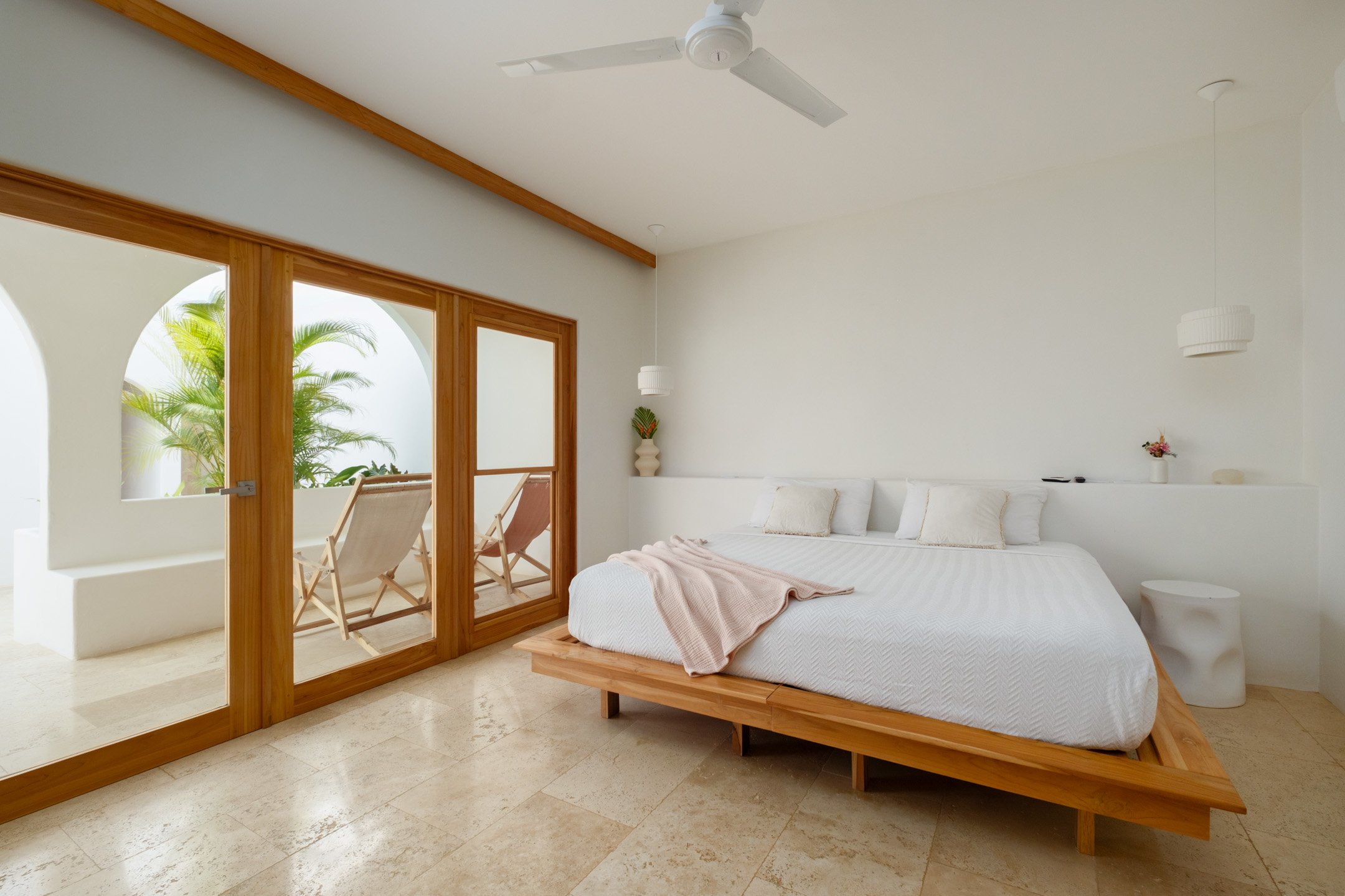 A bright, airy bedroom with white walls, a white bed with pillows and a blanket, and wood accents. There is a glass sliding door leading to a balcony with two deck chairs and potted plants outside.