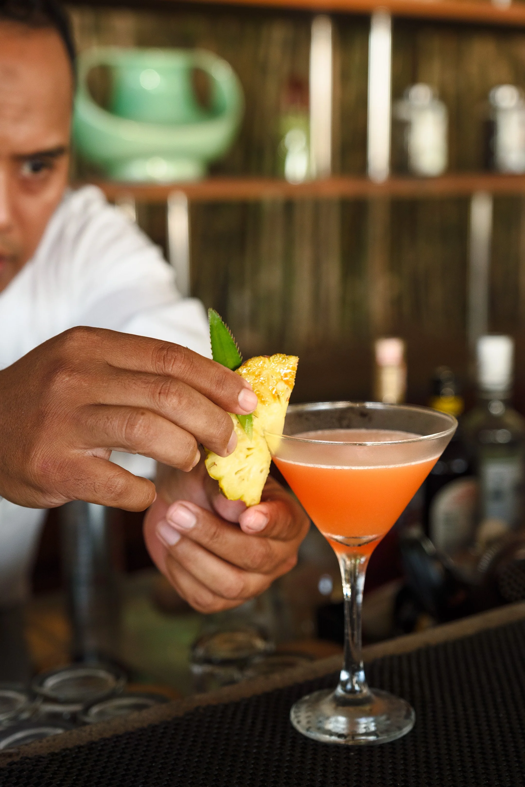 A bartender is garnishing a pink cocktail with a pineapple wedge and a lime slice in a martini glass.