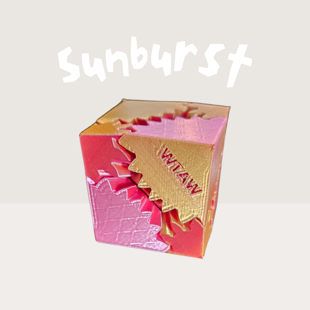 Colorful 3D-printed gear fidget cube in gold, pink, and red shades, labeled "WTAW", featuring an intricate rotating gear design, named the Sunburst edition for sensory stimulation and fidgeting.