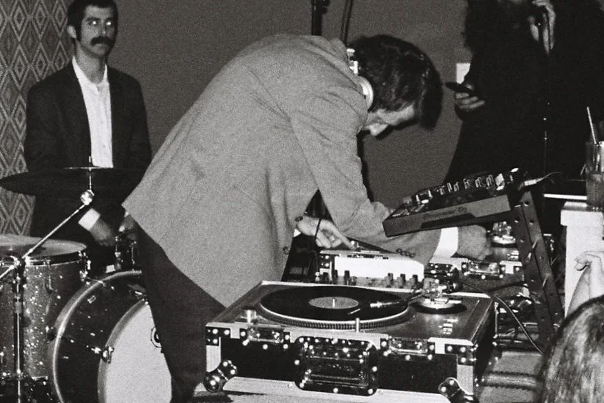 DJ wearing headphones and a suit, working with turntables and a mixer, with a drum set and audience in the background.