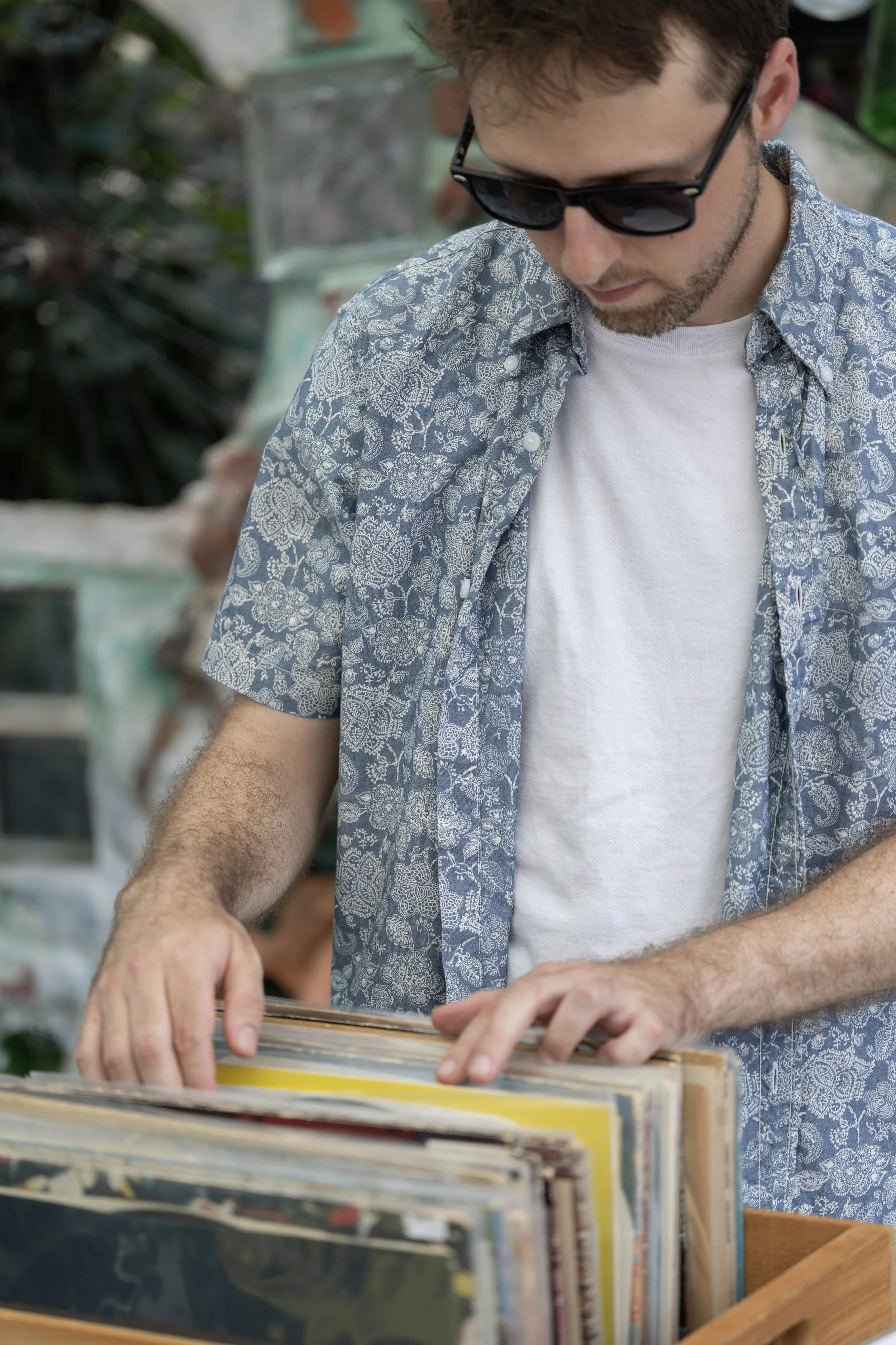 A man wearing sunglasses, a white t-shirt, and an open blue floral shirt is browsing through vinyl records in an outdoor setting.
